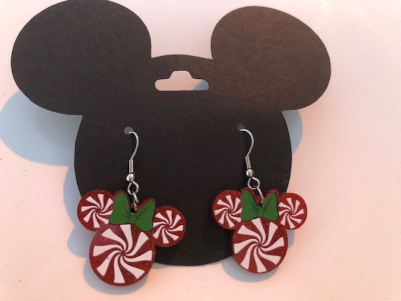 May include: A pair of red and white peppermint candy cane themed dangle earrings with green bows. The earrings are shaped like Mickey Mouse ears.