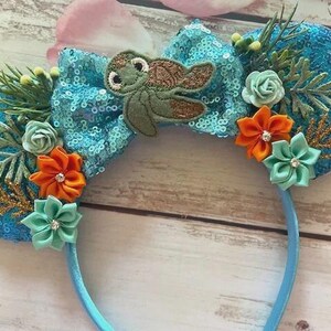 May include: A whimsical headband featuring teal sequined ears adorned with orange and teal fabric flowers, faux greenery, and a teal sequined bow with a turtle applique. The headband is light blue.