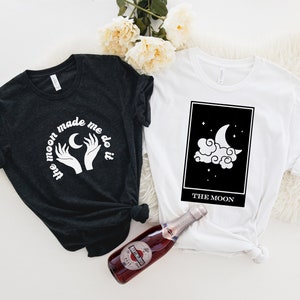 May include: Two t-shirts featuring a moon and stars design. One shirt is black with white text that reads "The moon made me do it". The other shirt is white with a black tarot card design that says "The Moon".