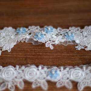 May include: Two white lace garters featuring intricate floral patterns and delicate scalloped edges. Each garter is accented with small, light blue fabric flowers, each centred with a tiny white pearl bead, creating a detailed and textured appearance.
