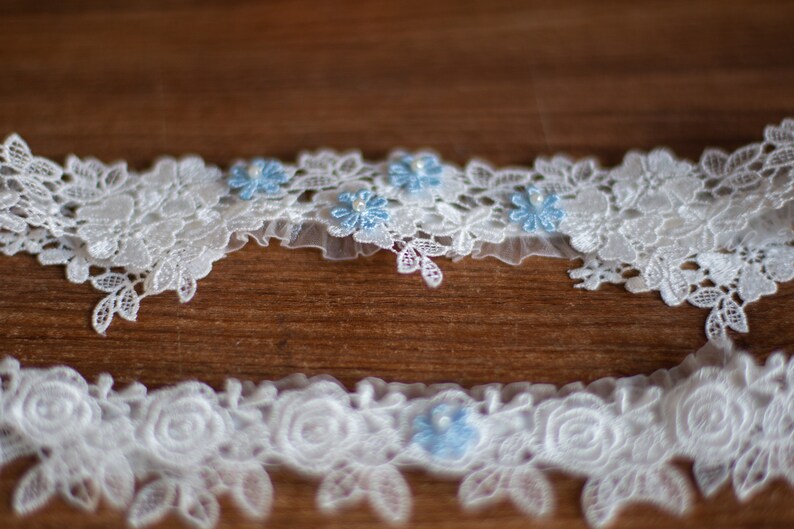 May include: Two white lace garters featuring intricate floral patterns and delicate scalloped edges. Each garter is accented with small, light blue fabric flowers, each centred with a tiny white pearl bead, creating a detailed and textured appearance.