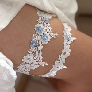 May include: A set of two white lace garters featuring intricate floral patterns and small light blue fabric flowers with central white beads. The wider garter displays a detailed lace design, while the thinner garter is accented with smaller floral motifs.