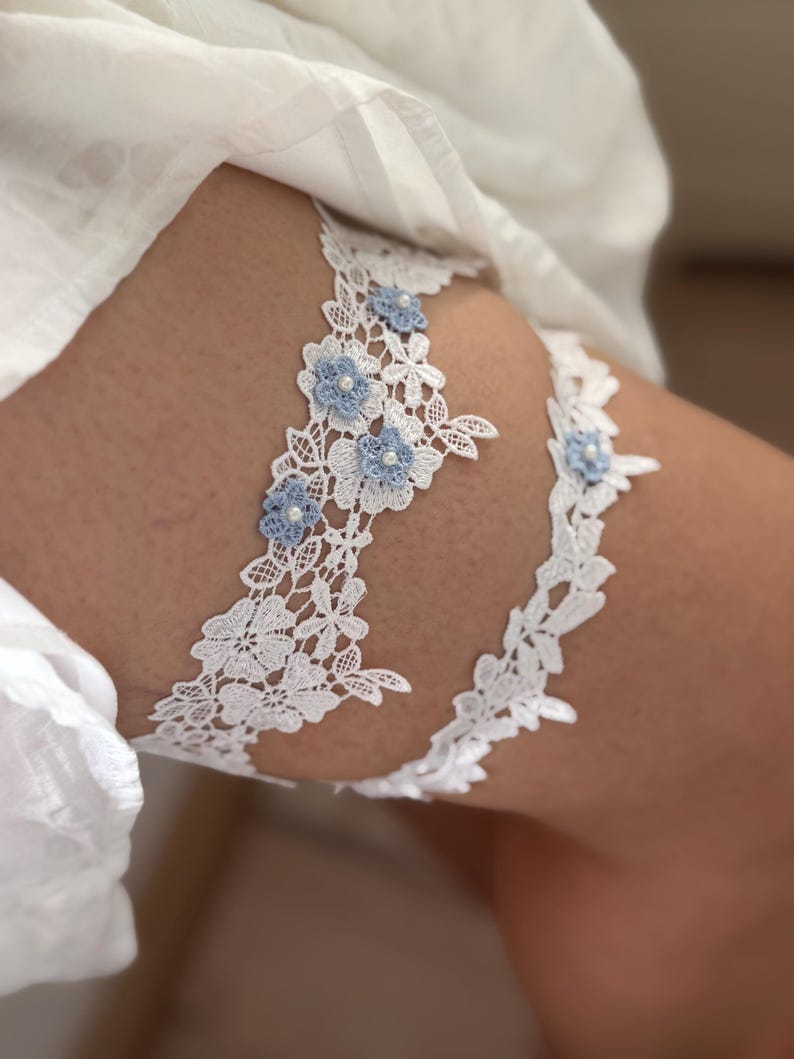 May include: A set of two white lace garters featuring intricate floral patterns and small light blue fabric flowers with central white beads. The wider garter displays a detailed lace design, while the thinner garter is accented with smaller floral motifs.