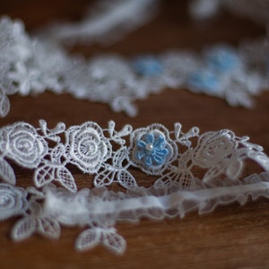 May include: A white lace trim featuring an intricate floral rose pattern with small light blue flower accents and a single white bead at the centre of each blue flower. The lace has a scalloped edge and a gathered white fabric ribbon base.
