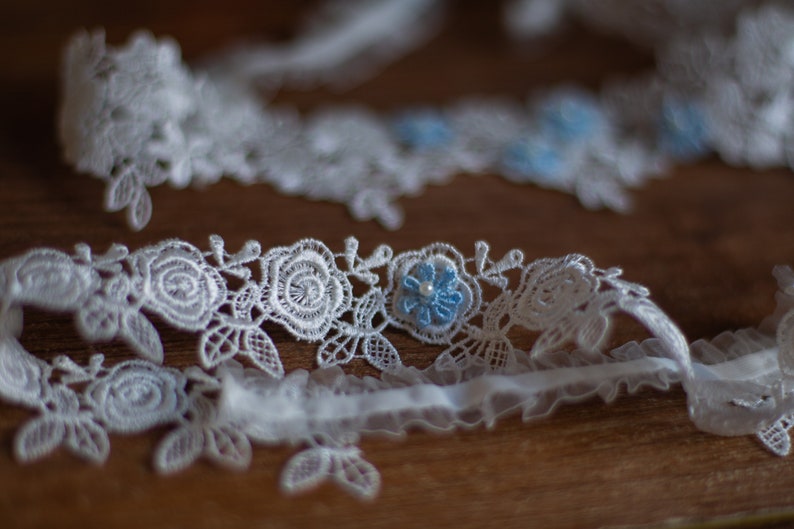 May include: A white lace trim featuring an intricate floral rose pattern with small light blue flower accents and a single white bead at the centre of each blue flower. The lace has a scalloped edge and a gathered white fabric ribbon base.