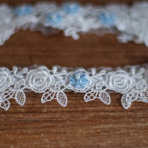 May include: A white lace garter featuring a repeating pattern of embroidered roses and leaves. The design is accented with small, light blue floral embellishments, each centred with a tiny white pearl bead, creating a delicate and intricate texture.