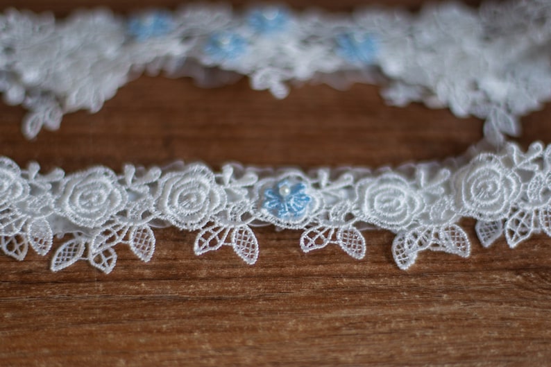 May include: A white lace garter featuring a repeating pattern of embroidered roses and leaves. The design is accented with small, light blue floral embellishments, each centred with a tiny white pearl bead, creating a delicate and intricate texture.