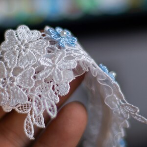 May include: A white lace trim featuring an intricate floral pattern with scalloped edges. Small, light blue fabric flowers are attached to the lace, each accented with a single, centred white pearl bead, creating a delicate and textured decorative border.