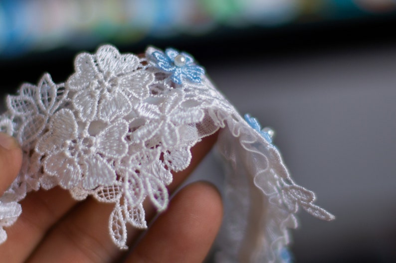 May include: A white lace trim featuring an intricate floral pattern with scalloped edges. Small, light blue fabric flowers are attached to the lace, each accented with a single, centred white pearl bead, creating a delicate and textured decorative border.