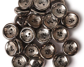 May include: A collection of round, metallic silver-toned buttons, each featuring a smooth, raised rim and a textured, recessed centre with two central holes for attachment. The buttons have a reflective, polished finish and are arranged in a cluster.