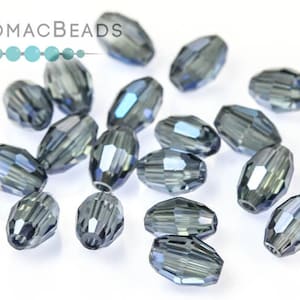 May include: A selection of faceted, oval-shaped beads in a smoky grey colour with iridescent highlights. The beads are scattered on a white surface. The PotomacBeads logo is in the upper left corner.