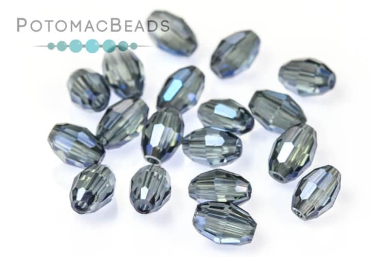 May include: A selection of faceted, oval-shaped beads in a smoky grey colour with iridescent highlights. The beads are scattered on a white surface. The PotomacBeads logo is in the upper left corner.