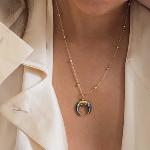 May include: A gold chain necklace with a crescent moon pendant made of iridescent abalone shell. The necklace is worn by a person with a white shirt.
