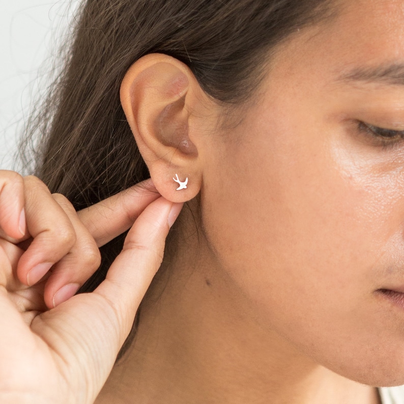 May include: A pair of silver stud earrings featuring a delicate, minimalist design of a small bird in flight.