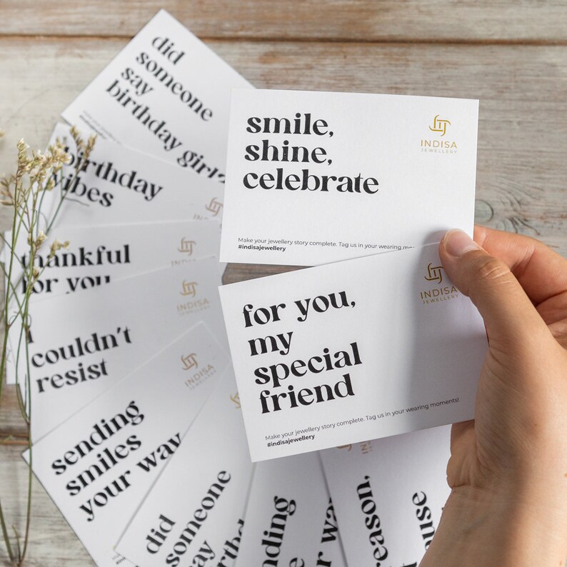 May include: A set of white cards with black text, each card featuring a different inspirational message. The cards are arranged in a pile, with the top card reading "smile, shine, celebrate". The cards are from the brand Indisa Jewellery.