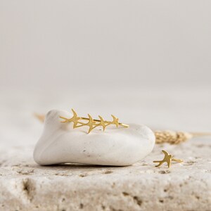 May include: A pair of gold earrings featuring a flock of birds in flight. The earrings are displayed on a white rock with a natural background.