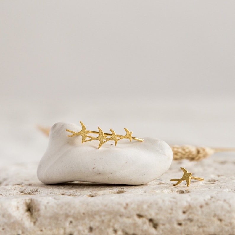 May include: A pair of gold earrings featuring a flock of birds in flight. The earrings are displayed on a white rock with a natural background.