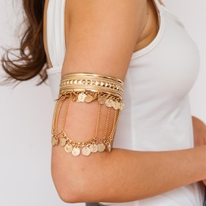 May include: Gold-coloured arm cuff with multiple bands and dangling coin charms. The upper band has a geometric design. The arm cuff is worn on the upper arm. The jewellery is a bohemian style.