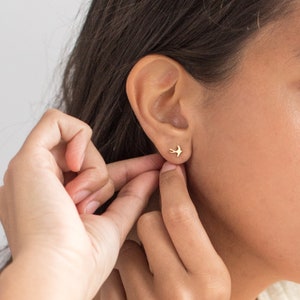 May include: A close-up of a person's ear with a small gold bird stud earring. The earring is simple and delicate, with a minimalist design.