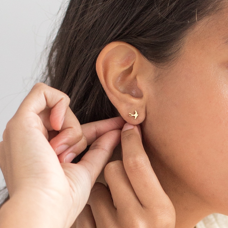 May include: A close-up of a person's ear with a small gold bird stud earring. The earring is simple and delicate, with a minimalist design.
