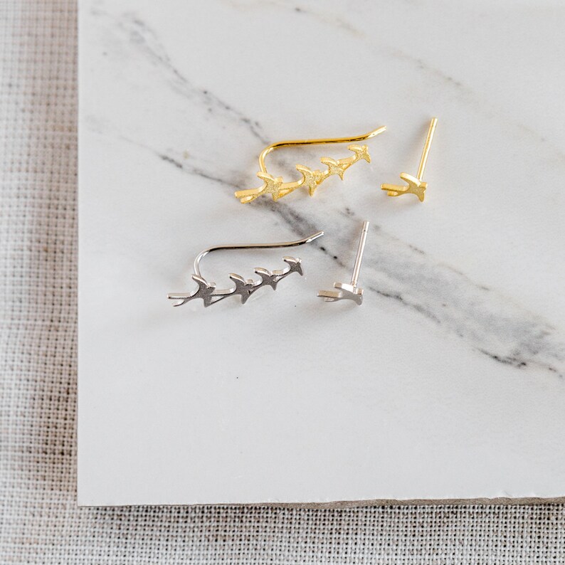 May include: Two pairs of ear climbers, one gold and one silver, each featuring a row of five small bird silhouettes.