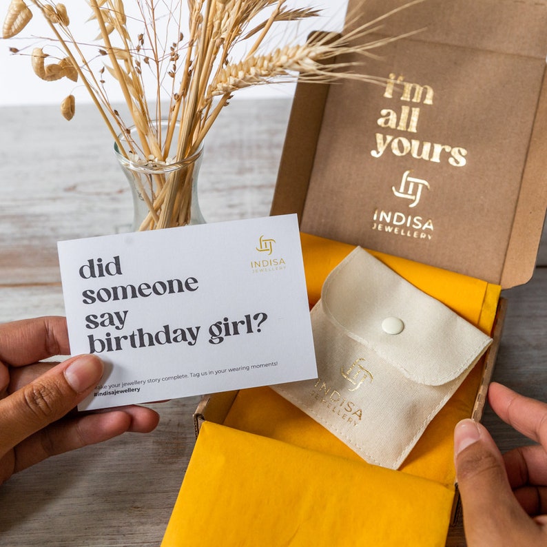 May include: A white card with the text "did someone say birthday girl?" and the Indisa Jewellery logo. The card is held by a hand. The card is in front of a brown box with the text "I'm all yours" and the Indisa Jewellery logo. The box is open and has a yellow lining. A small white pouch with the Indisa Jewellery logo is inside the box.