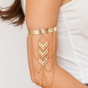 May include: Gold arm cuff with a textured band and dangling chain details. The cuff features three chevron-shaped pendants and multiple delicate chains, creating a layered effect. The jewelry is worn on the upper arm.