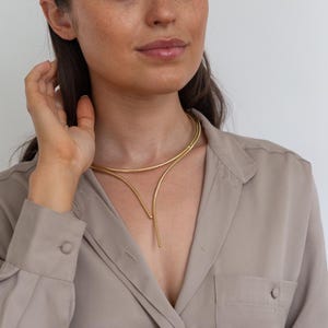 May include: A gold-toned double-layered necklace with a curved design. The necklace has a long, thin pendant that hangs down the front.