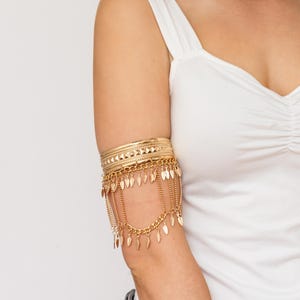 May include: Gold-coloured arm cuff with multiple bands and dangling leaf-shaped charms. The upper arm jewellery features a layered design with intricate detailing and chain accents, creating a bohemian aesthetic. The cuff is worn on the upper arm.