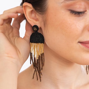 May include: Black and gold beaded earrings with a fringe design. The earrings are long and dangle from the earlobe.