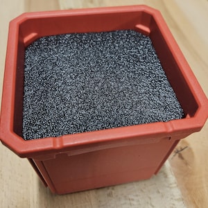 May include: A red plastic planter filled with black, granular material. The planter has a square shape with slightly angled sides and a textured surface. The black material fills the planter to the top, creating a contrasting color and texture.