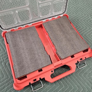 May include: Open, red plastic tool case with a clear lid. The case has a handle and metal latches. Inside are two gray foam inserts. The top of the case has several small compartments. The brand name "Milwaukee" is visible on the lid.