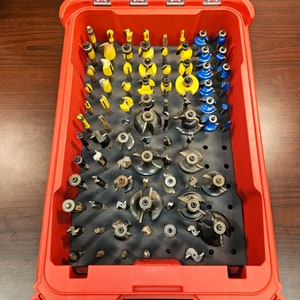 May include: A red plastic storage case with a clear lid, filled with a variety of router bits. The bits are organized in rows and columns, with different sizes and shapes. The case has a handle on the top and a latch on the front.