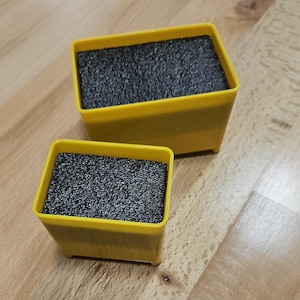 May include: Two yellow rectangular containers with a dark gray, textured foam insert. The containers are different sizes and are placed on a light brown wooden surface. The containers are empty.