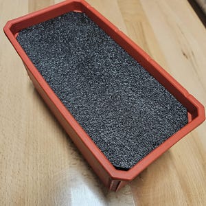 May include: A rectangular, red plastic storage container with a textured, dark gray foam insert. The container has a slightly beveled edge and sits on a light wood surface. The container is empty and ready for use.