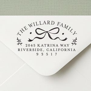 May include: A white envelope with a black stamp featuring the text "THE WILLARD FAMILY" and address details. The stamp includes a bow design and decorative elements. The envelope is surrounded by greenery and light blue ribbon.