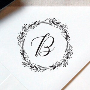 May include: A black and white illustration of a wreath with leaves and berries surrounding the letter "B".