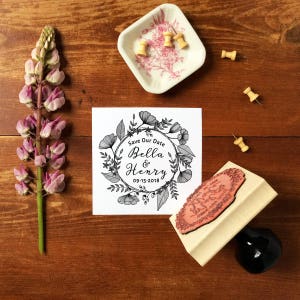 Puede incluir: Una tarjeta blanca con un dise&ntilde;o de corona floral en blanco y negro. El texto "Save Our Date Bella & Henry 09-15-2018" est&aacute; dentro de la corona. Un sello de madera con un dise&ntilde;o floral est&aacute; al lado de la tarjeta.