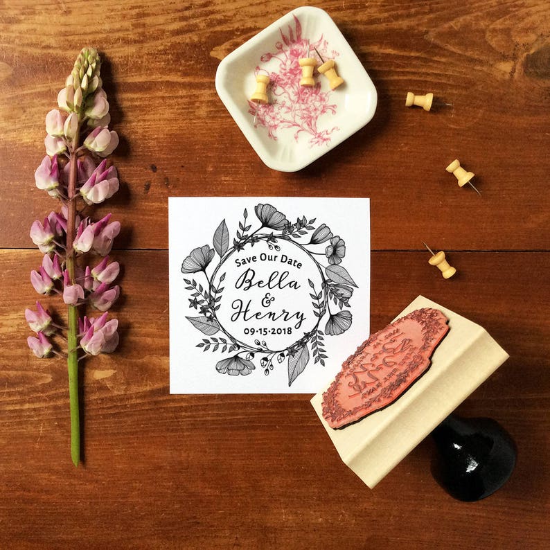 Puede incluir: Una tarjeta blanca con un dise&ntilde;o de corona floral en blanco y negro. El texto "Save Our Date Bella & Henry 09-15-2018" est&aacute; dentro de la corona. Un sello de madera con un dise&ntilde;o floral est&aacute; al lado de la tarjeta.