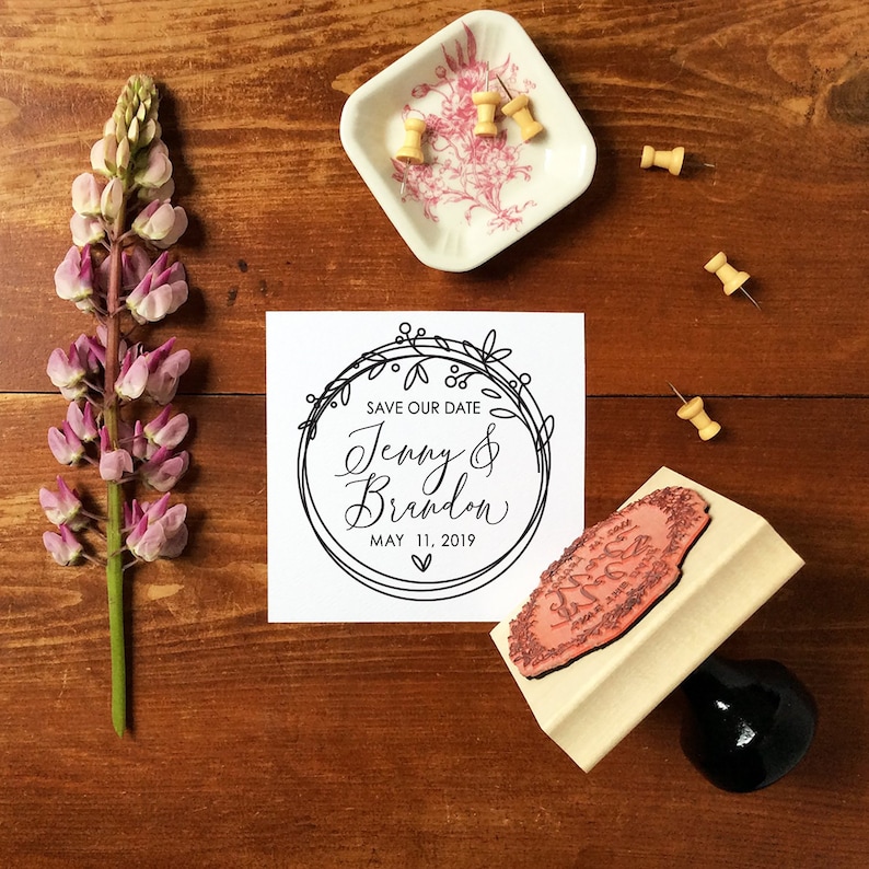 以下が含まれることがあります： 「Save Our Date Jenny & Brandon May 11, 2019」というテキストが書かれた、白地に黒と白のリースのイラストが描かれた白いカードです。