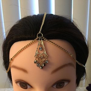 Peut inclure: Tiare de tête en chaîne dorée avec un pendentif en cristal. Le pendentif est de forme triangulaire avec un motif en filigrane et de petits cristaux.