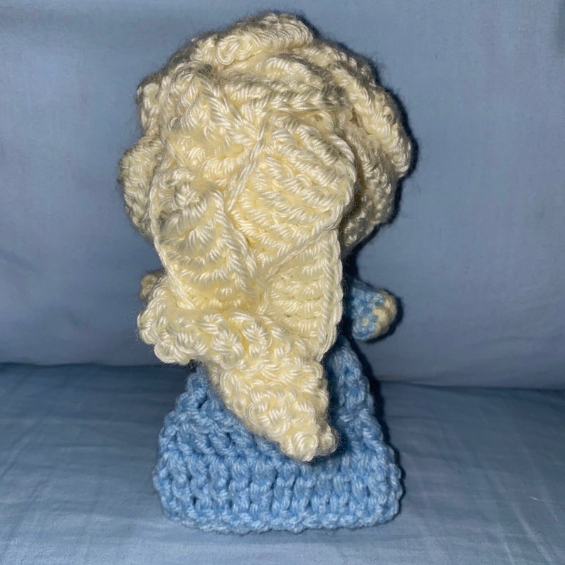 May include: A crocheted doll with a light blue dress and a cream-colored crocheted hat. The doll is sitting on a blue surface.
