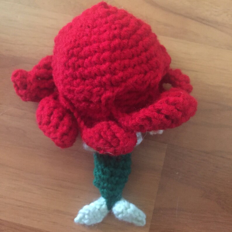 May include: A handmade crochet mermaid doll with a large red head and flowing hair. The mermaid has a green tail and white fins. The doll is made of yarn and is sitting on a wooden surface.