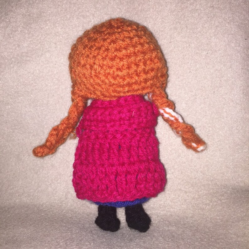 May include: A crocheted doll with orange hair, a pink dress, and blue shoes. The doll is standing on a white surface.