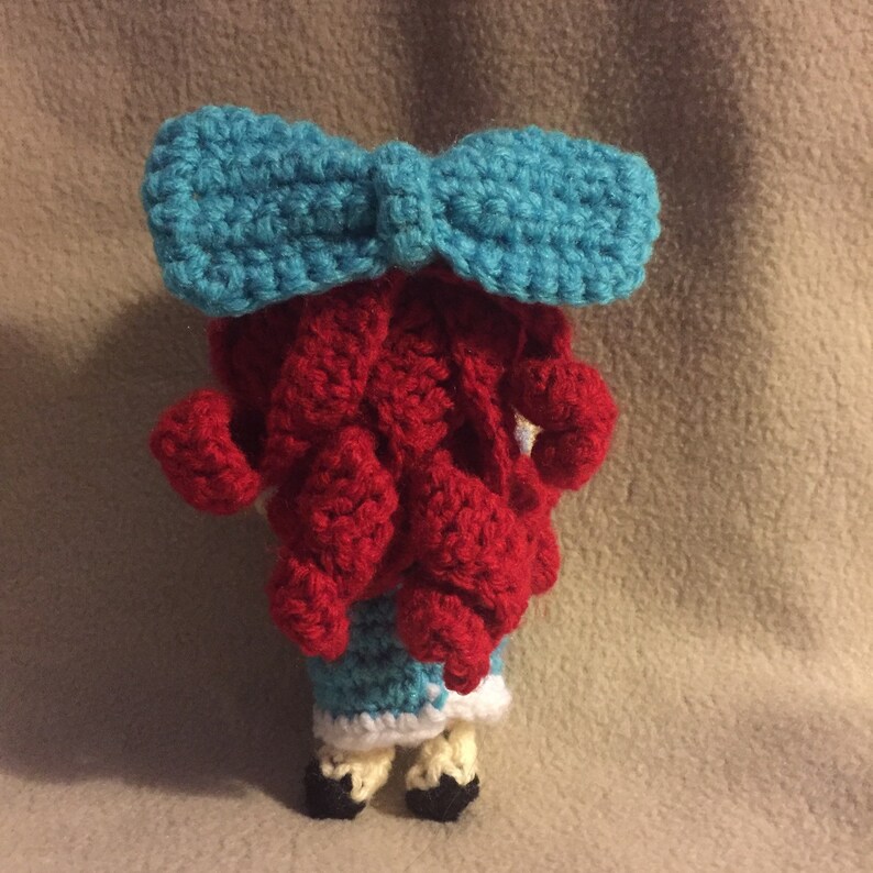 Puede incluir: Una mu&ntilde;eca de crochet con pelo rojo, un lazo azul y un vestido azul y blanco. La mu&ntilde;eca lleva zapatos negros.