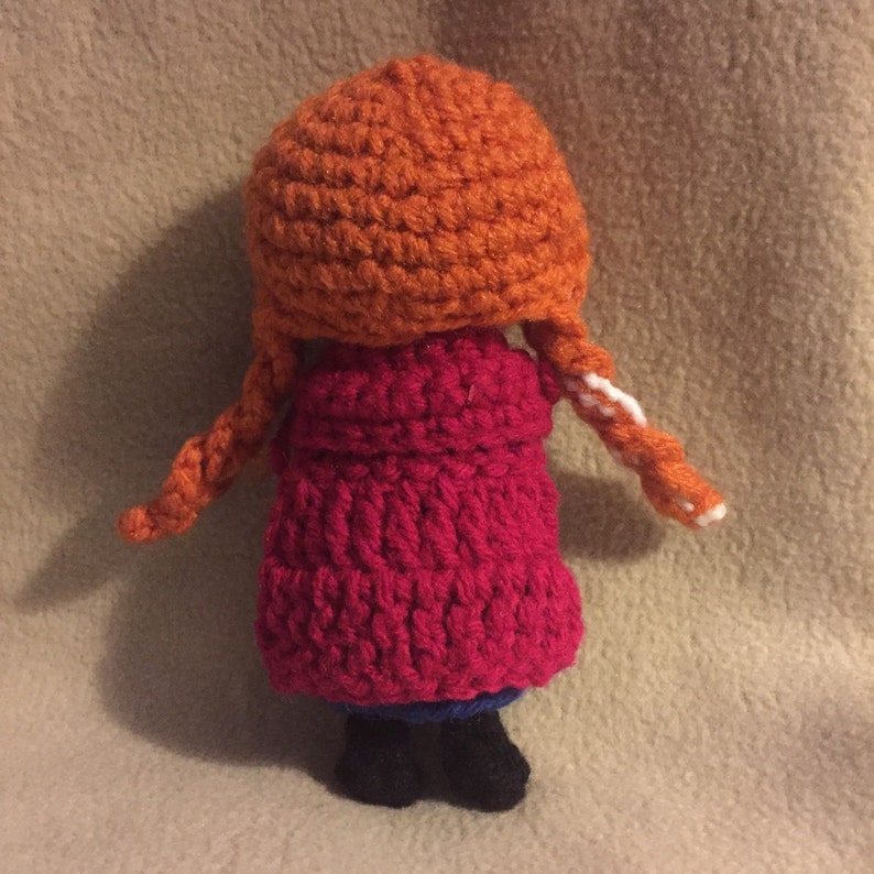 May include: A crocheted doll with red hair, a red coat, and black shoes. The doll is wearing a white and orange hat.