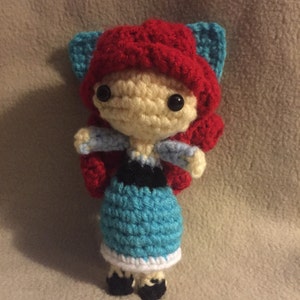 Puede incluir: Una mu&ntilde;eca de crochet con pelo rojo, un vestido azul y blanco y zapatos negros. La mu&ntilde;eca tiene una cara de color amarillo claro con ojos negros y un gorro de crochet rojo con orejas de gato azules.