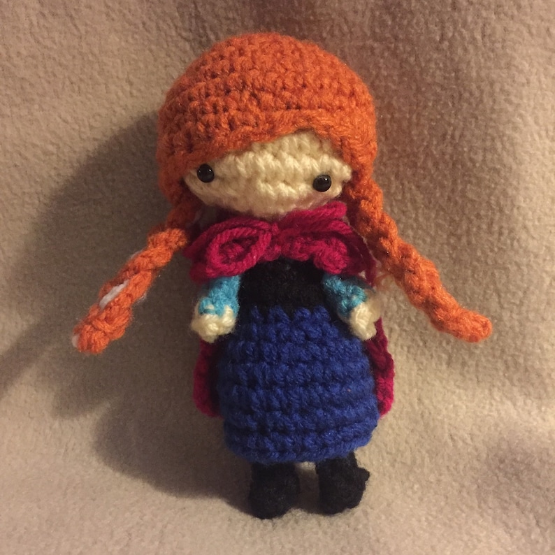 May include: A crocheted doll with red hair, a blue dress, and a red cape. The doll has a white face with black eyes and a red bow.