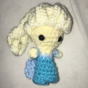 May include: A crocheted doll with blonde hair, a blue dress, and black eyes. The doll is sitting on a white surface.