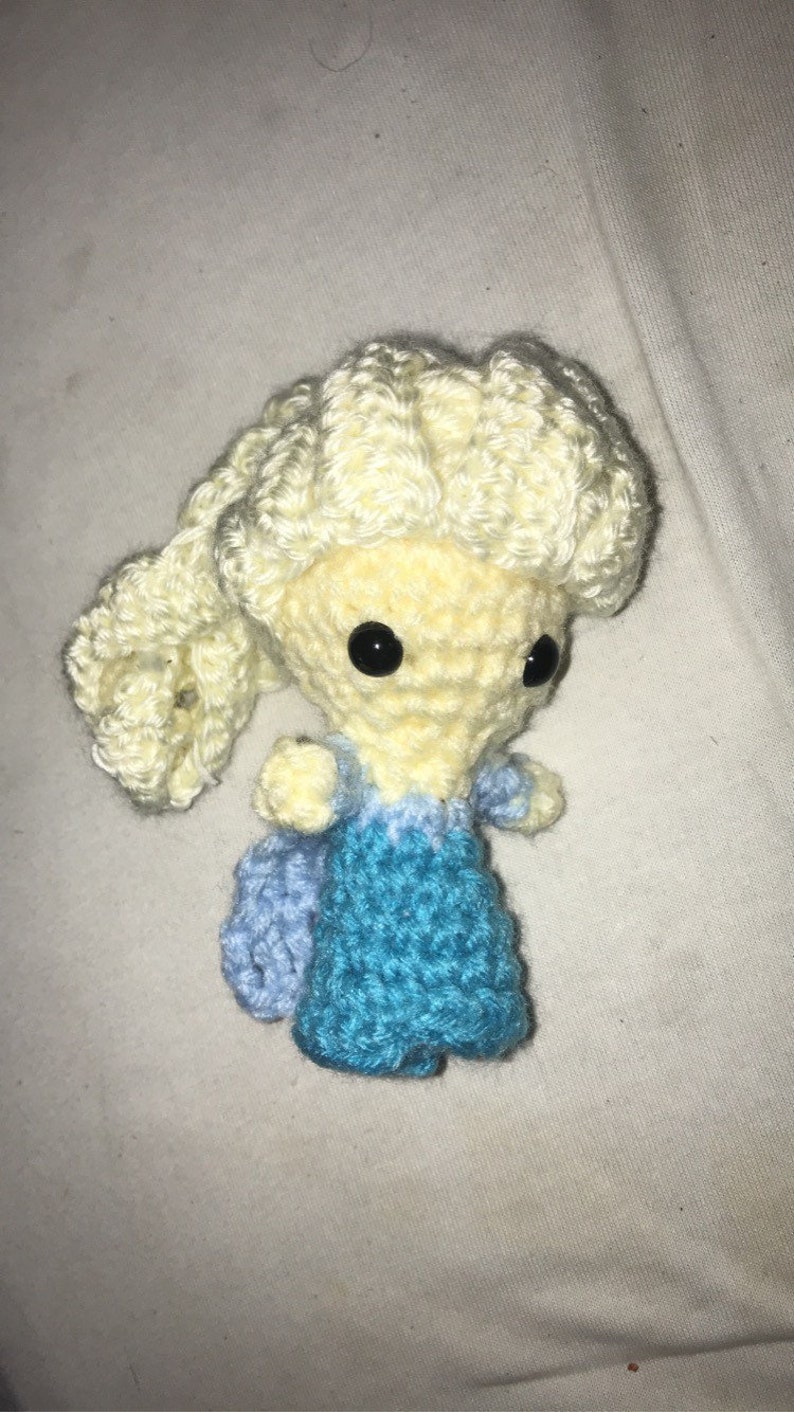 May include: A crocheted doll with blonde hair, a blue dress, and black eyes. The doll is sitting on a white surface.